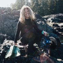 Agathe sitting on a rocky shore, holding filming and scuba gear. End of image description.