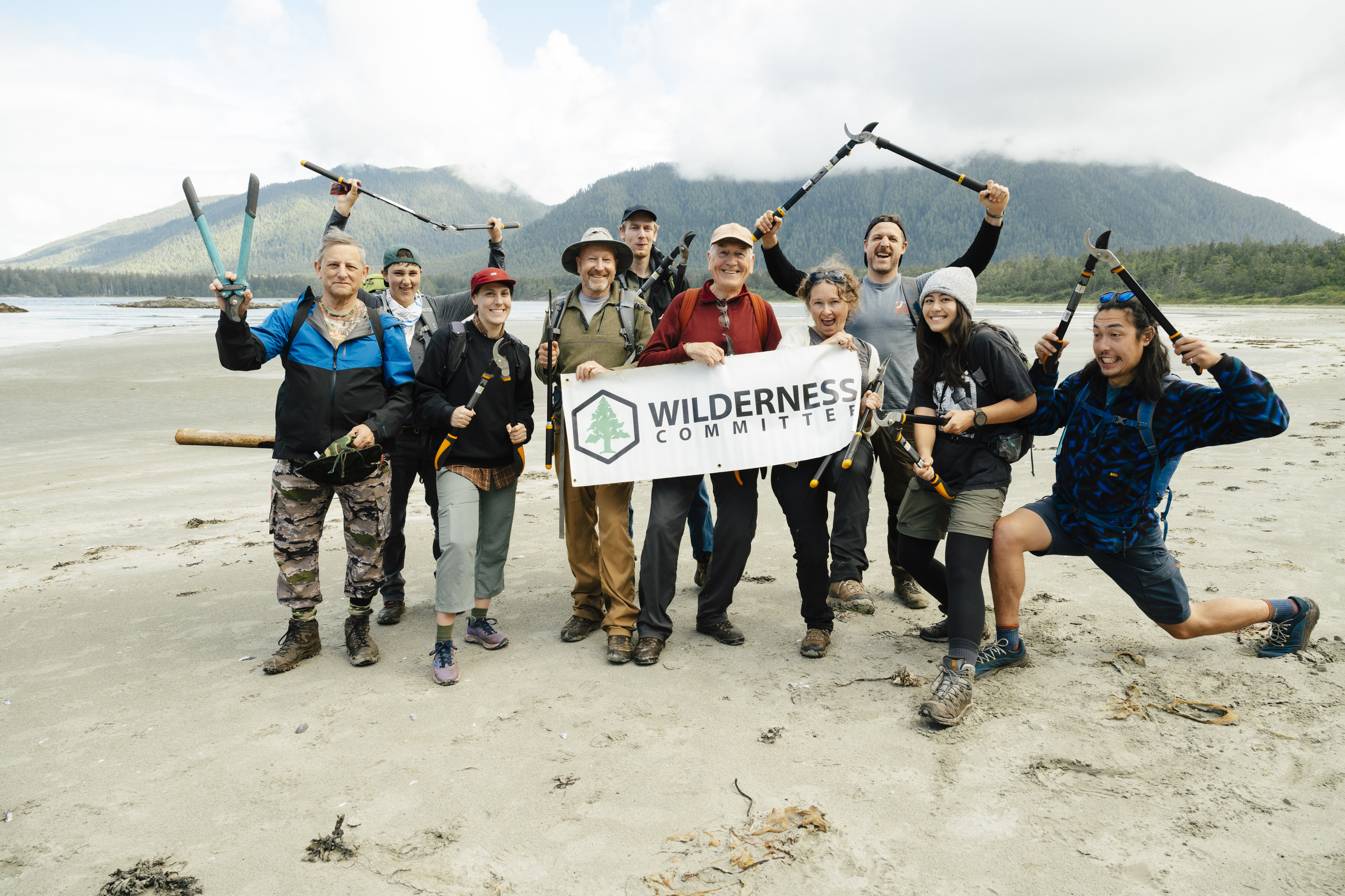 A group of people on a beach with trail building tools in their hands and a sign that reads Wilderness Committee. End of image description.