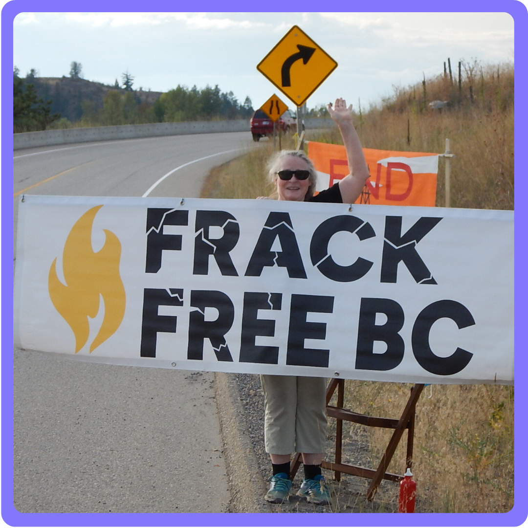 A woman standing at the side of a highway holding a Frack Free BC banner. end of image description.