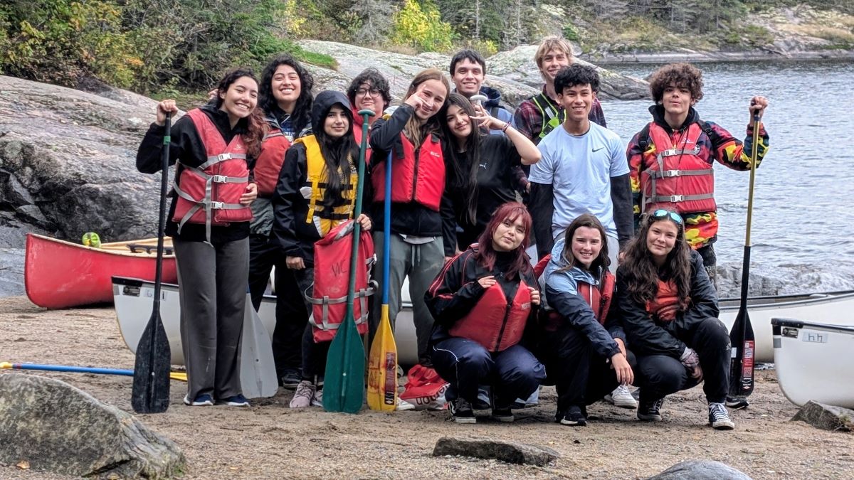 A group of teenagers by a river with life jackets and paddles. end of image description.