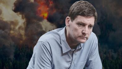 B.C. premier David Eby frowning at the camera while a wildfire burns behind him. End of image description.