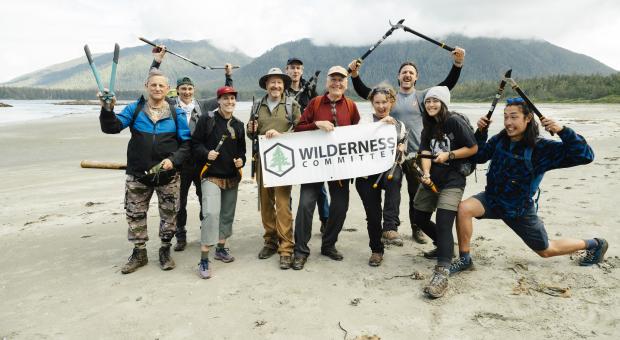 A group of people on a beach with trail building tools in their hands and a sign that reads Wilderness Committee. End of image description.