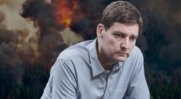 B.C. premier David Eby frowning at the camera while a wildfire burns behind him. End of image description.